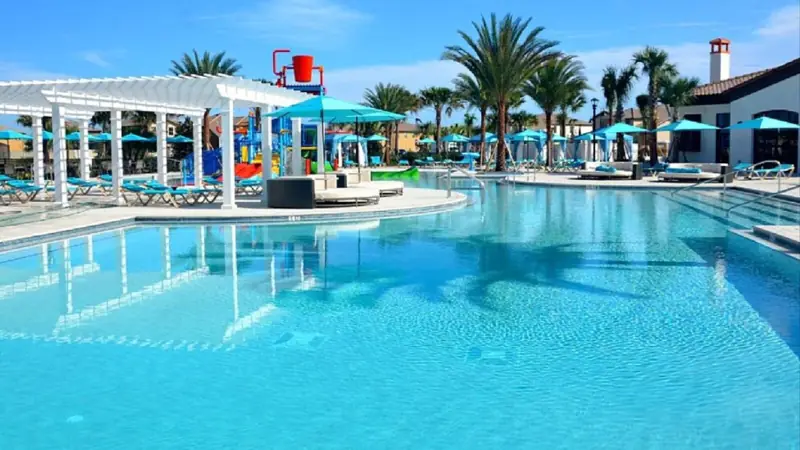 Clubhouse pool – A swimming pool with lounge chairs and umbrellas, surrounded by palm trees and a covered area with a water slide.